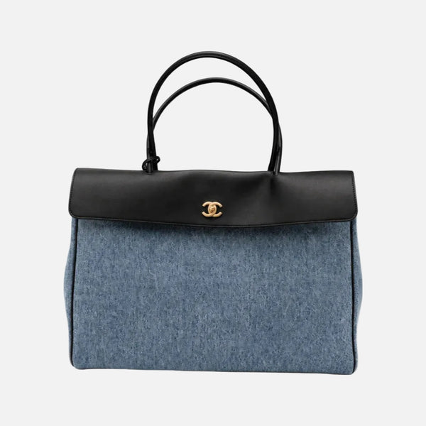 A Chanel handbag featuring a blue denim body, a black leather top flap and handles, and a gold-tone interlocking CC logo on the front, set against a plain light gray background.