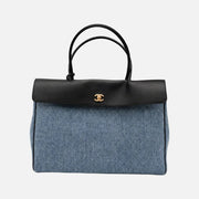 A Chanel handbag featuring a blue denim body, a black leather top flap and handles, and a gold-tone interlocking CC logo on the front, set against a plain light gray background.