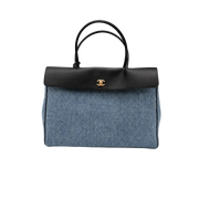 A Chanel handbag featuring a blue denim body, a black leather top flap and handles, and a gold-tone interlocking CC logo on the front, set against a plain light gray background.